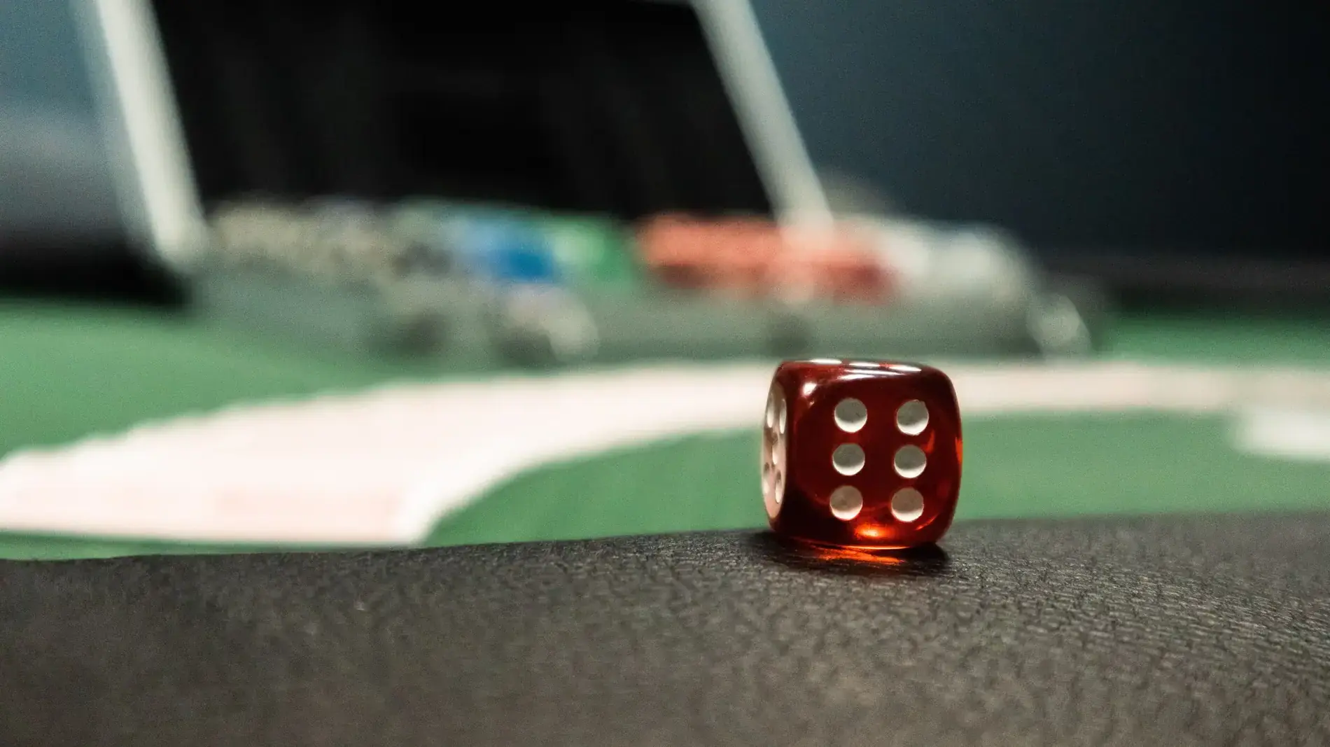 A red die on the edge of a casino table, with chips and playing cards blurred behind it.